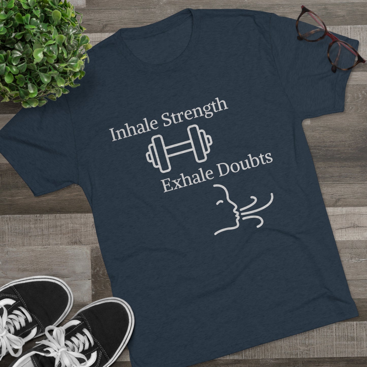 A motivational t-shirt in gray with the Inhale Strength Exhale Doubt w Images - Unisex Tri-Blend Crew Tee by Printify displayed on a wooden floor, accompanied by black sneakers and a pair of glasses.