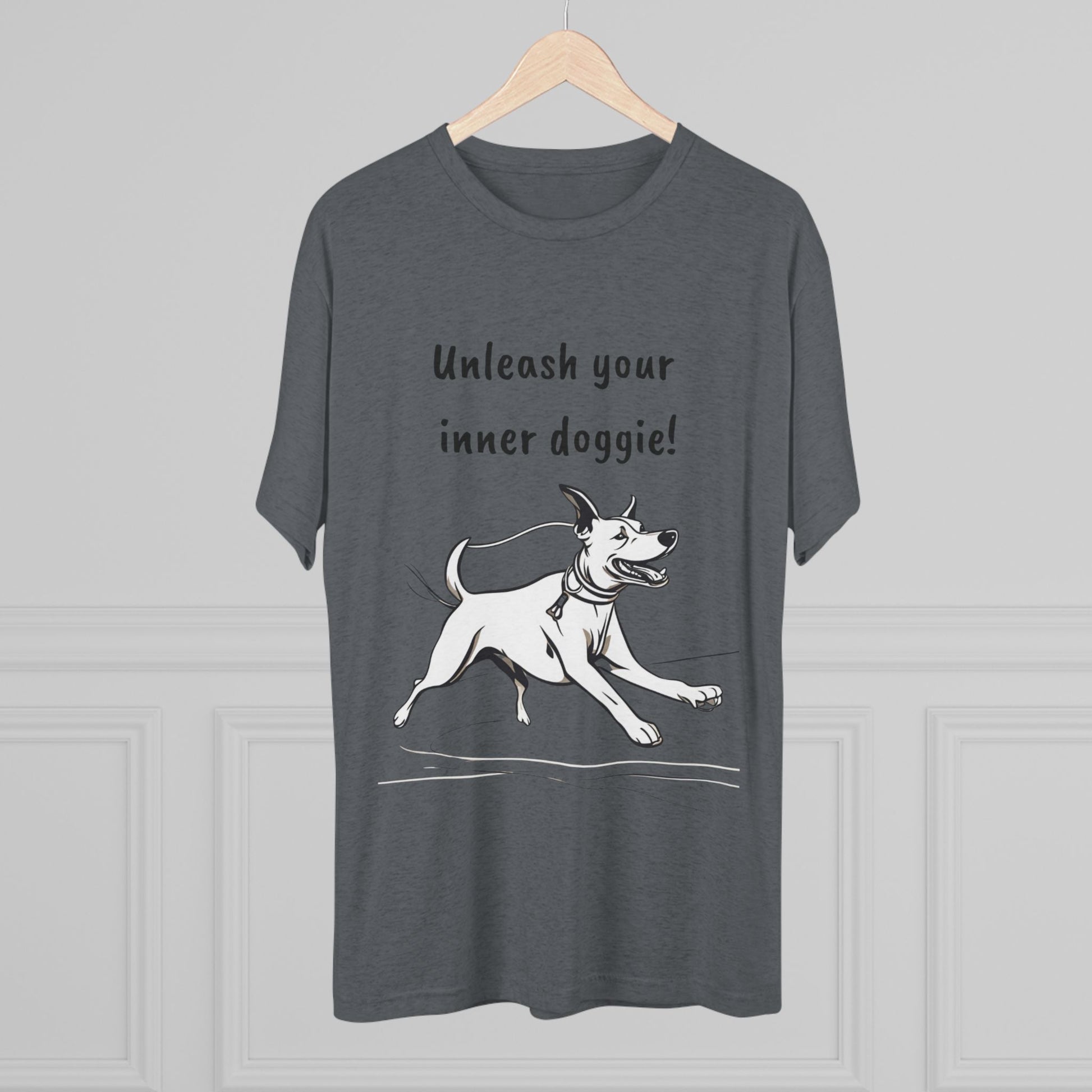 The "Unleash your inner doggie" Tri-Blend Crew Tee by Printify, hanging on a wooden hanger, showcases a white cartoon dog running with its tongue out and a joyful expression on a dark grey fabric. The whimsical design includes the text "Unleash your inner doggie!" above the playful pup. Made from a premium blend of fabrics for versatile fashion, this unisex fit tee is displayed against a light grey wall with subtle panel detailing.