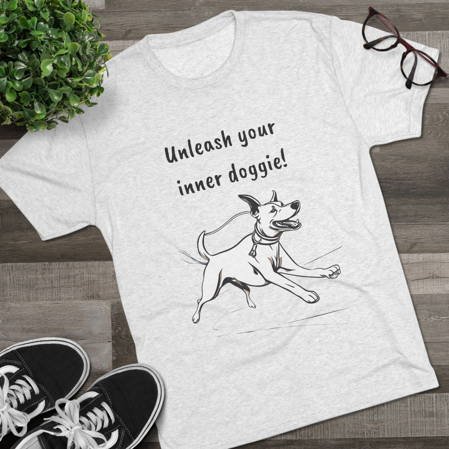 A light gray *Unleash your inner doggie* Tri-Blend Crew Tee in a unisex fit from Printify, featuring a premium blend and a black-and-white illustration of a playful dog, is displayed on a wooden floor next to a pair of black sneakers, eyeglasses, and a leafy plant. The shirt is printed with the text "Unleash your inner doggie!" using sustainable practices.