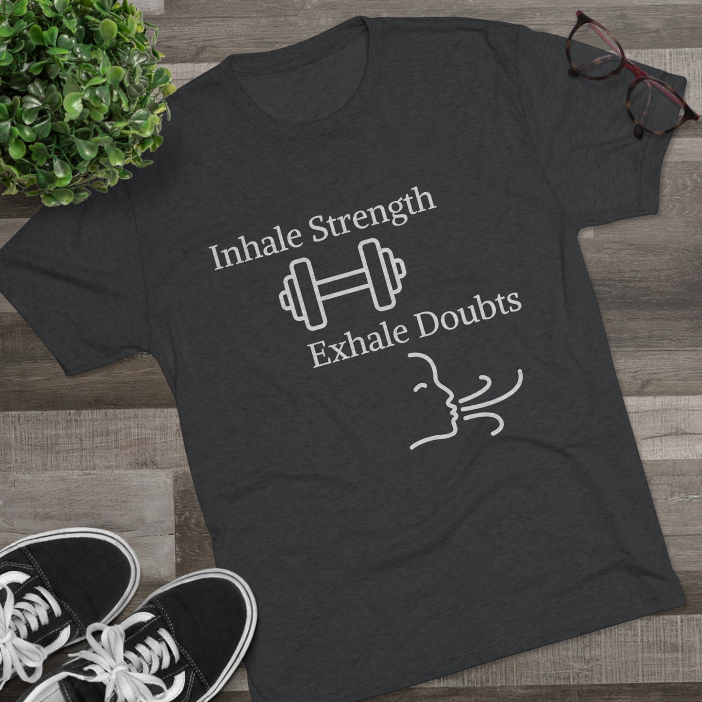 A gray motivational Printify t-shirt with the phrase "Inhale Strength Exhale Doubt w Images" and a graphic of a dumbbell and swirls, laid flat on a wooden surface alongside sneakers and glasses.