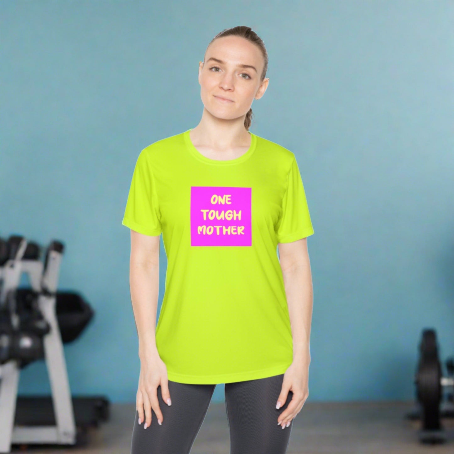 A woman in a gym wearing a bright green Printify Ladies Competitor Tee - One Tough Mother with the phrase "one tough mother" in bold pink and white text. She stands confidently, posing with her hands on her hips.