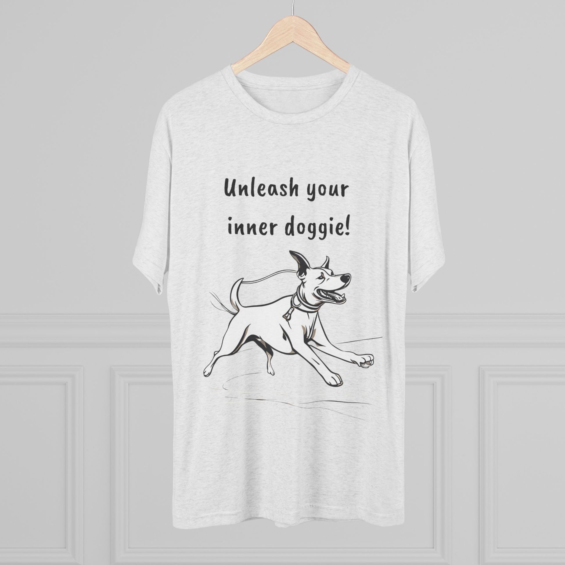 A Unleash your inner doggie - Tri-Blend Crew Tee from Printify is showcased on a wooden hanger against a light gray background. The premium blend shirt features a black-and-white design of a joyful dog running, with the text "Unleash your inner doggie!" printed above the illustration.