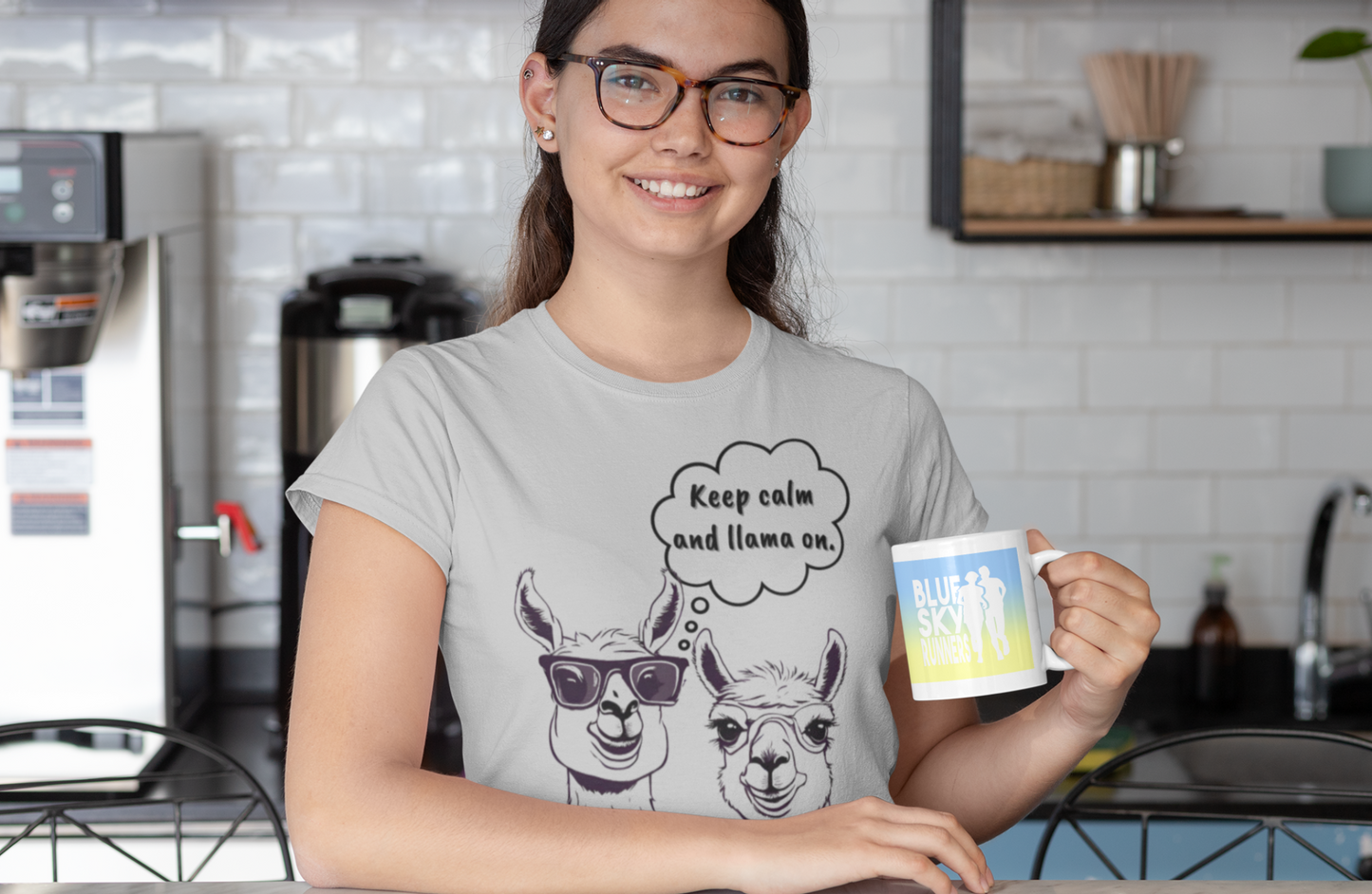 A woman wearing glasses and a comfortable grey Printify Keep Calm and Llama On - Tri-Blend Crew Tee - Unisex Fit smiles while holding a mug that says "Blue Sky Coffee." She stands in a modern kitchen with white subway tiles and coffee-making equipment in the background.