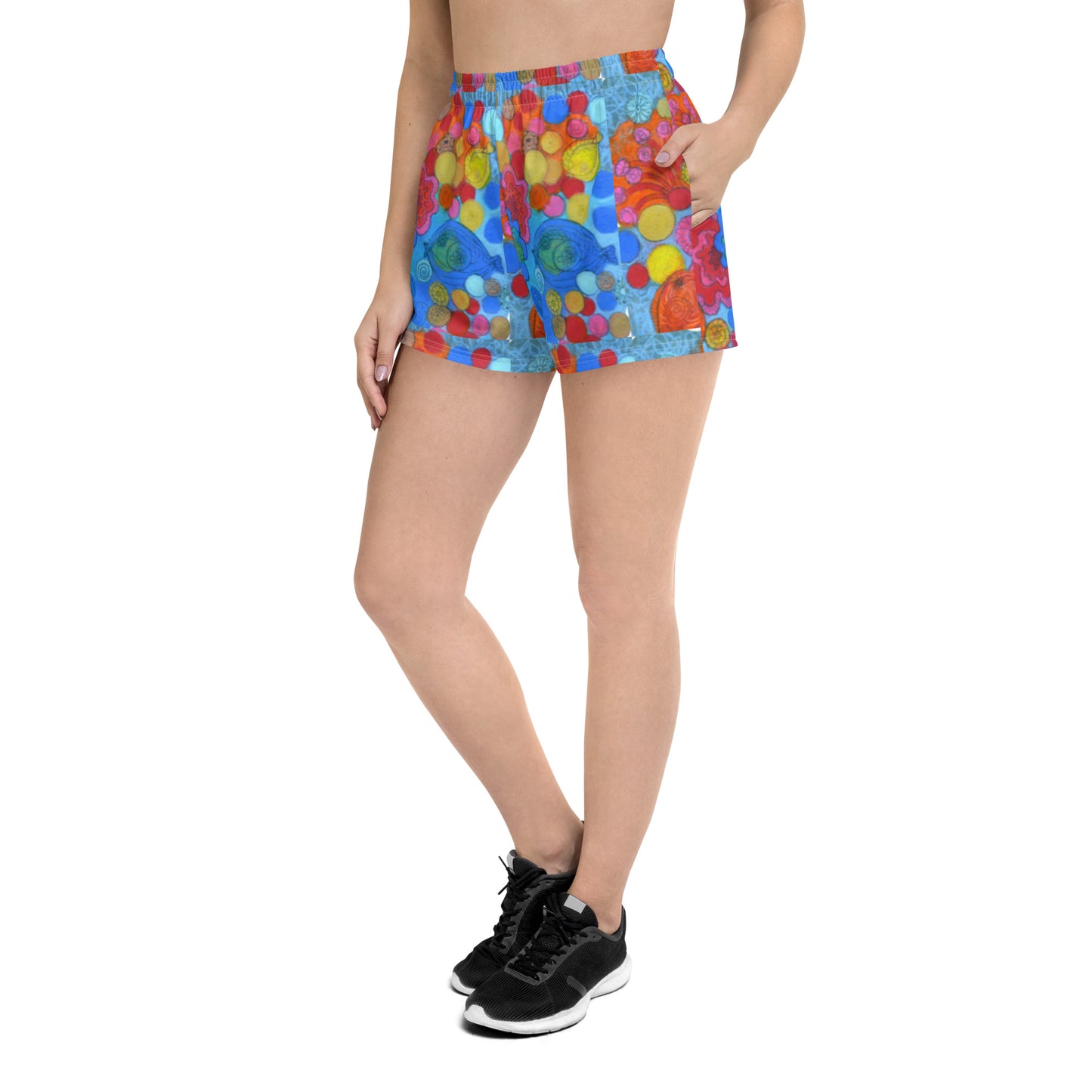 A person is confidently standing with one hand in their pocket, wearing Blue Sky Runners' Coral - Women's Recycled Athletic Shorts. These vibrant shorts feature various shapes and colors such as red, yellow, and green on a blue background. The outfit is completed with a black T-shirt and black sneakers.