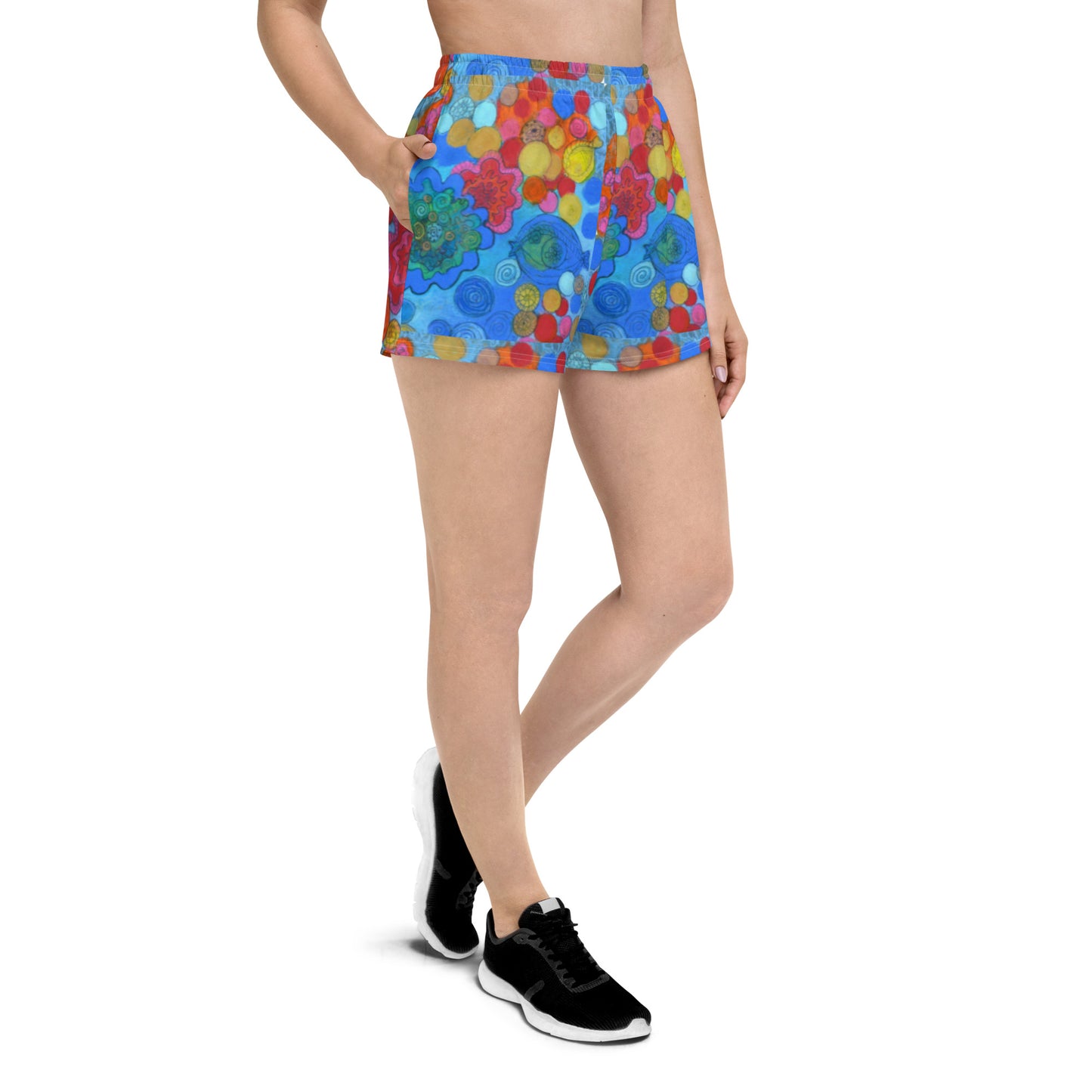 A person is standing with one hand in the pocket of their Blue Sky Runners Coral - Women’s Recycled Athletic Shorts, which are adorned with a vibrant, multicolored coral print pattern featuring flowers and circles. They are wearing a white top and black athletic shoes. The background is plain and white.