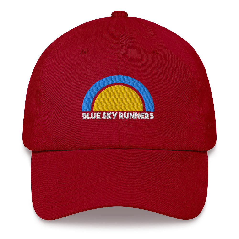 The Blue Sky Runners - Dad Hat by Blue Sky Runners is a red low profile hat with an embroidered semicircle in blue and yellow. Below the semicircle, "BLUE SKY RUNNERS" is embroidered in white. The hat also features an adjustable strap for a perfect fit.