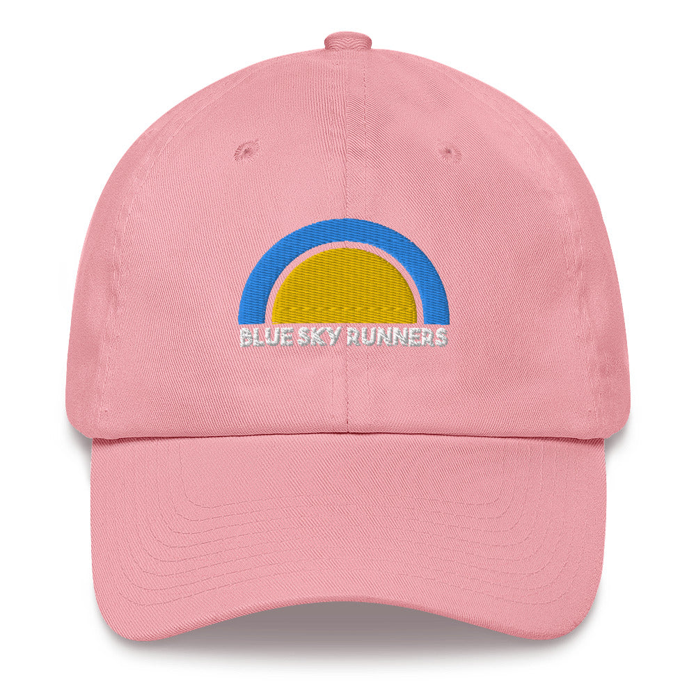 The Blue Sky Runners - Dad Hat is a low profile pink baseball cap featuring an embroidered design on the front. The design includes a half-circle rainbow in blue, yellow, and white, with the text "BLUE SKY RUNNERS" written in white below the rainbow. It also comes with an adjustable strap for a perfect fit.